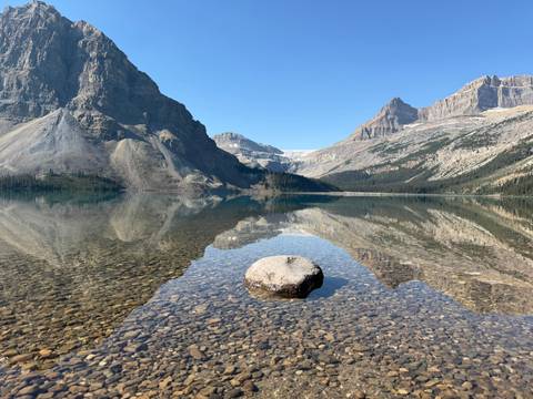 Crystal-clear alpine lake reflecting rugged mountains with single foreground rock