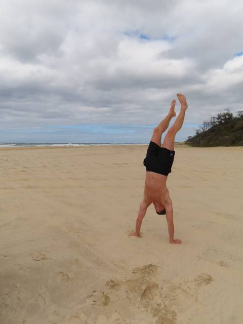 A shirtless man performs a handstand on a wide, empty sandy beach under cloudy skies.