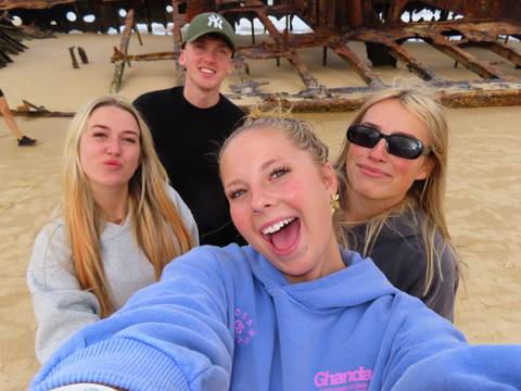 Friends take a cheerful selfie on a beach beside the rusted Maheno shipwreck.