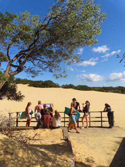 Travellers gather near a vast yellow sand dune under a bright blue sky.