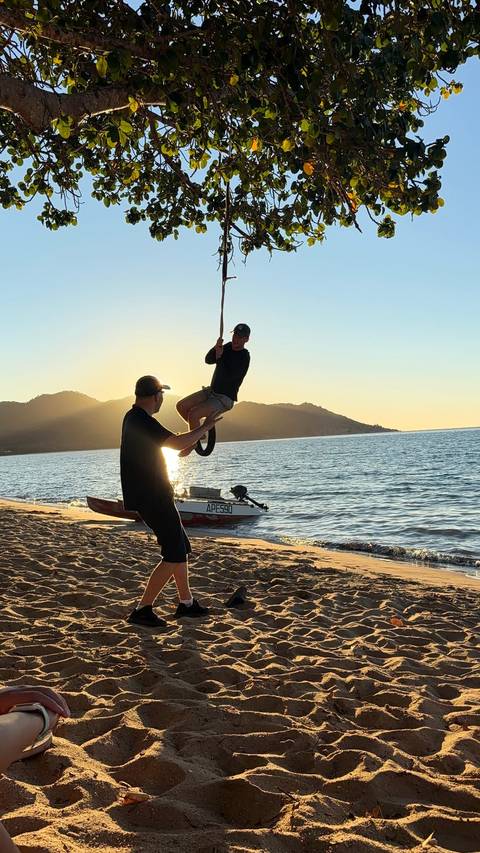 At sunset, a traveller swings from a rope over a sandy beach while a friend steadies the rope.