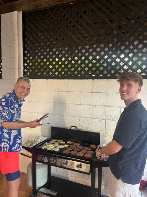 Two young men smile while preparing a barbecue in an outdoor communal area.