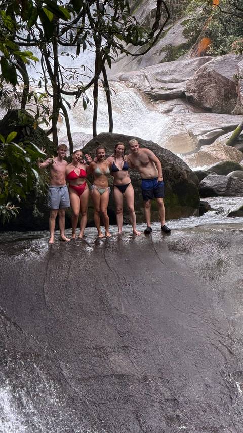 Travellers in swimwear stand on wet rocks beside a rushing freshwater stream in a rainforest gorge.