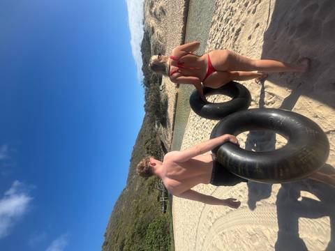 A man and woman carry large inner tubes down a sunny sandy beach toward a creek.