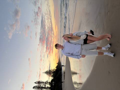 Couple pose on a tranquil beach at dawn with colourful clouds reflecting on wet sand.