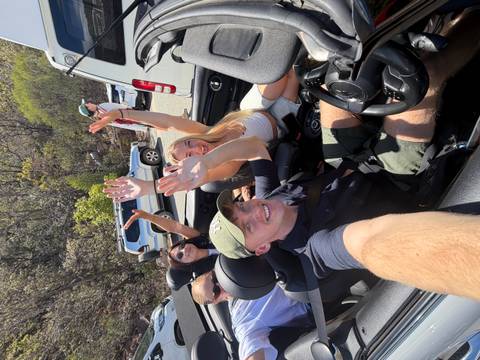 Group riding in a convertible cheer with raised arms on a sunny scenic road.