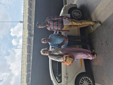 Two tourists pose with their driver beside a white taxi outside an airport terminal