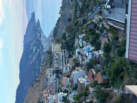 A coastal hillside town with terracotta rooftops cascades toward a curving blue bay backed by mountains.