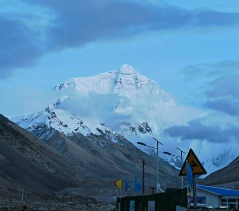Snow-clad Himalayan peak emerging through clouds above rugged foothills at dusk-blue hour.