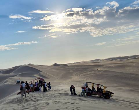 Tourists gather around sand-buggies on vast sun-lit dunes under a hazy afternoon sky.