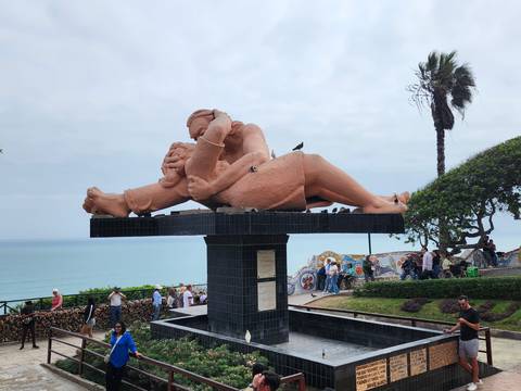 The iconic 'El Beso' sculpture of two lovers embraces above Lima’s seafront park with visitors around.