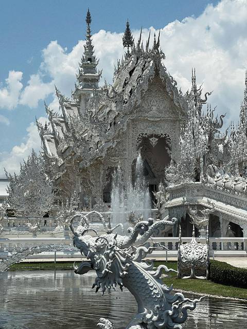 Intricately ornate white facade of Wat Rong Khun with fountains spraying in front.