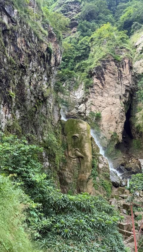 Tall rocky gorge with a thin waterfall cascading beside a large moss-covered elephant head sculpture.