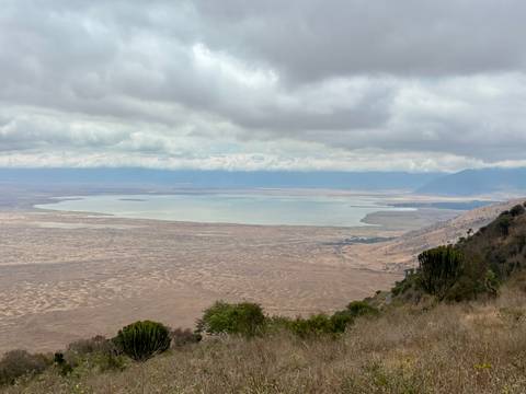 Expansive view from crater rim over a broad lake and patterned plains beneath a cloudy sky.