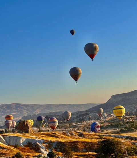 Hot-air balloons rise high above layered hills in the soft early morning blue sky.