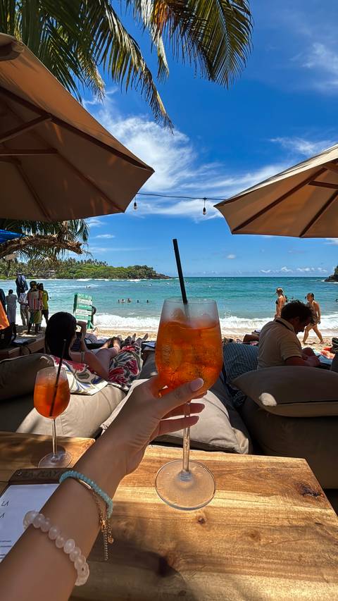 Beachgoers lounge under umbrellas while a hand holds up a chilled orange cocktail overlooking turquoise waves and a tropical shoreline.