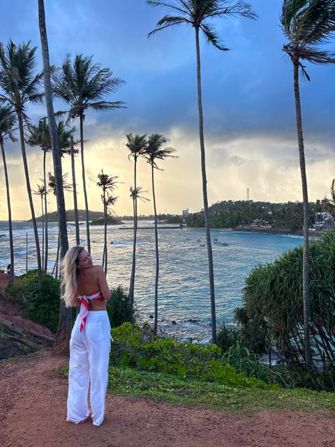 A woman poses on a windy clifftop lined with tall palm trees overlooking a choppy turquoise bay at sunset.