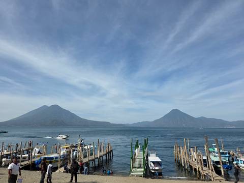 Lake Atitlán stretches between two imposing volcanic cones under streaked blue-white skies, with boats moored at a wooden jetty in the foreground.