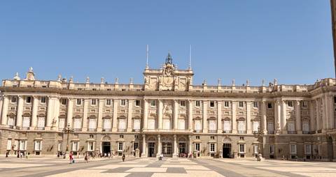 Wide façade of an ornate royal palace overlooking a spacious courtyard with scattered visitors.