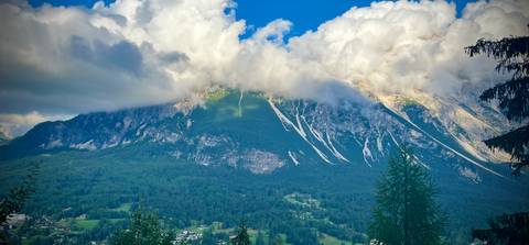 Dramatic Dolomite mountain partly covered by thick, low-lying clouds with green forest and villages below.