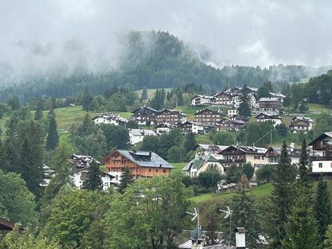 Misty alpine village with wooden chalets scattered across rolling green hills and conifer forests.