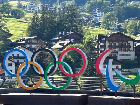 Colorful Olympic rings sculpture set against alpine village houses and green hills in bright sunlight.