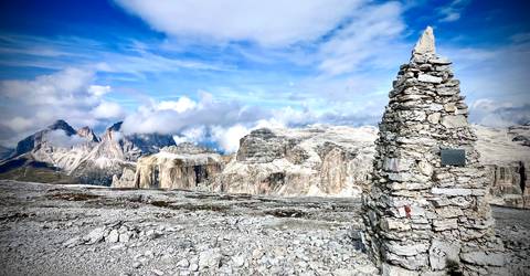 Rock-strewn high-altitude plateau in the Dolomites with a tall stone cairn foreground and dramatic rocky peaks beyond.