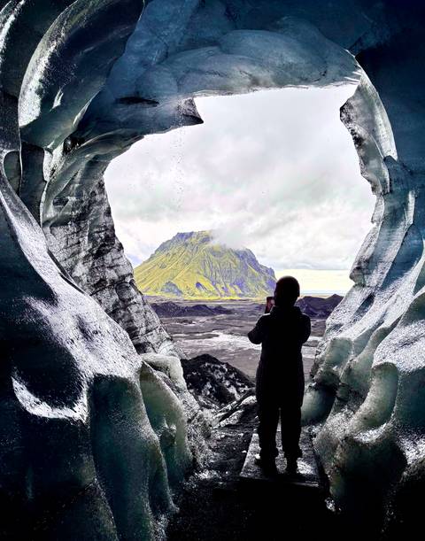 Silhouette of traveler framed by blue ice cave opening, photographing a moss-covered volcanic mountain.