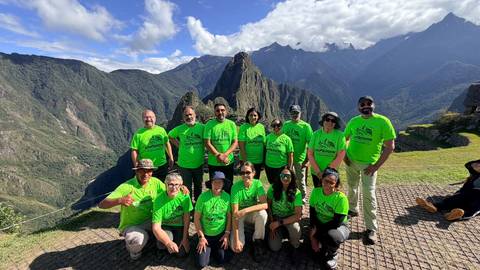 Large trekking group in matching green shirts posing with Machu Picchu citadel and Andean peaks behind.
