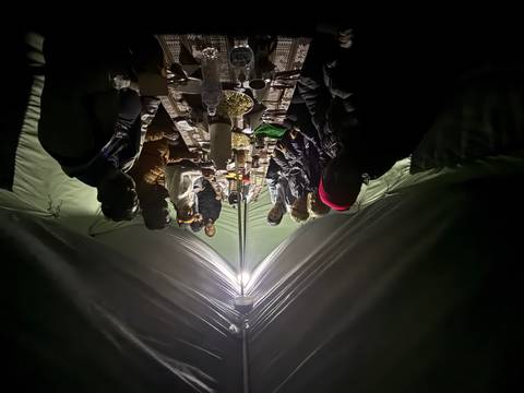 Night-time meal inside a camping tent, trekkers gathered around a long table lit by a central lamp.