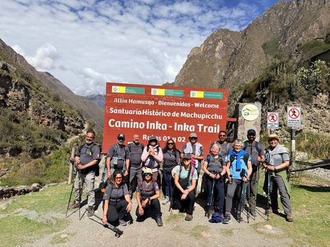 Full trekking group with poles posing at Inca Trail entrance sign in bright sunshine.