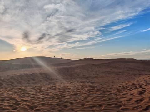Sun setting behind undulating Sahara dunes casting long rays and dramatic sky.
