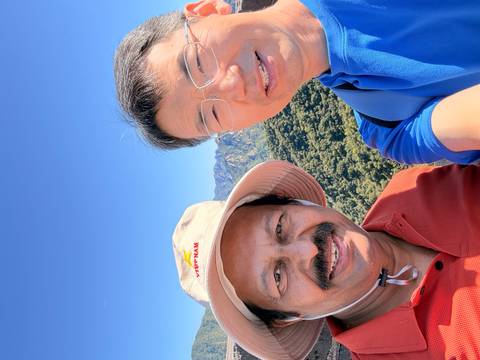 Two men taking a selfie with green forested mountains and clear blue sky in the background