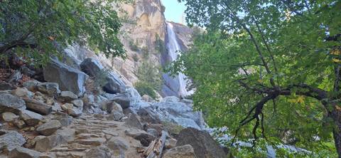 Rocky stair trail leading toward a cascading waterfall surrounded by granite cliffs and green trees in Yosemite.