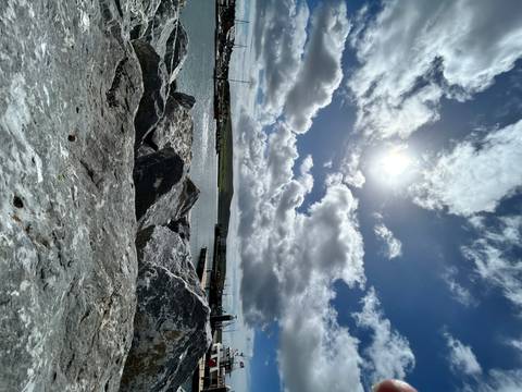 Rocky harbor foreground with bright sun shining through dramatic clouds over calm water and distant pier