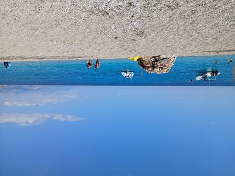 Pebble beach with clear blue Adriatic Sea, anchored boats and swimmers enjoying sunny weather.