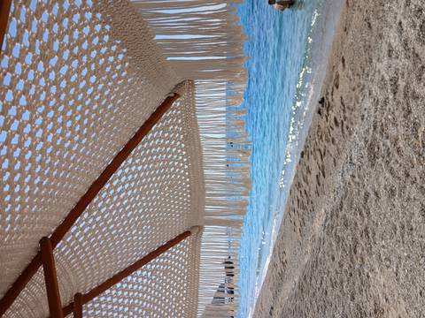 View of a sandy beach and calm sea framed by the fringe of a woven parasol.