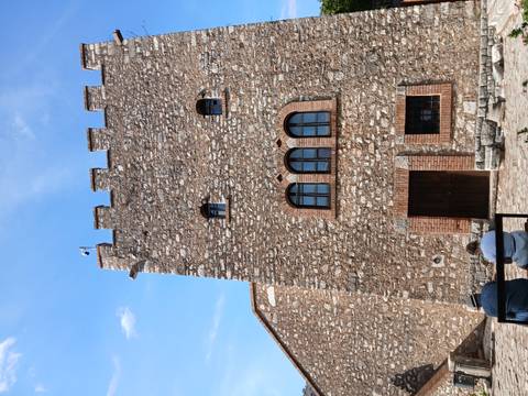 Stone fortress tower with small arched windows under a blue sky, two visitors seated in foreground.