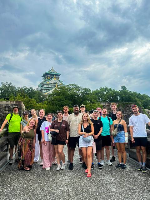 Tour group smiling on a stone bridge with Osaka Castle rising behind green trees