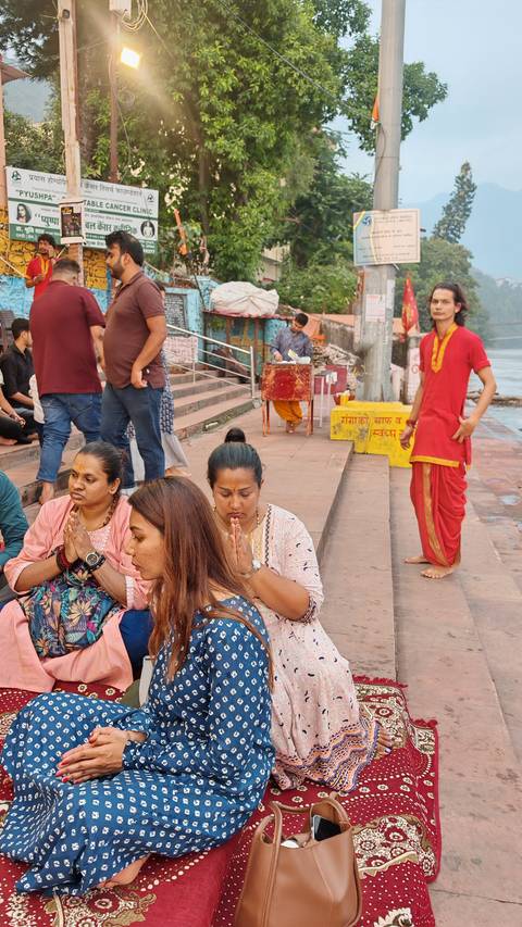 Women seated on ghats with hands folded in prayer while a priest prepares offerings nearby