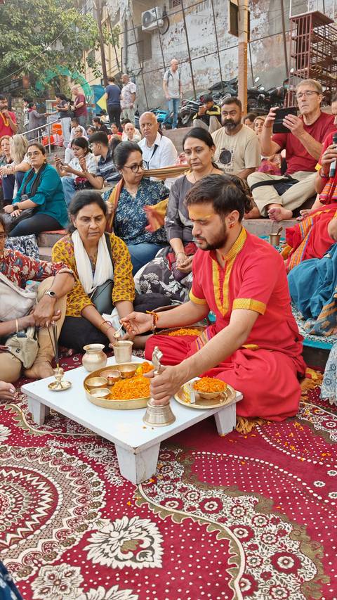 Hindu priest in red leading a ritual with brass vessels and marigold petals surrounded by devotees
