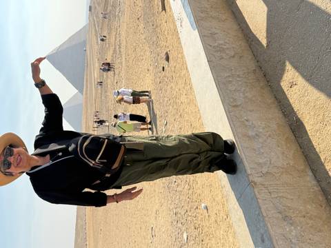 A smiling woman poses on a ledge, pinching the tip of two pyramids on the Giza Plateau under bright sun.