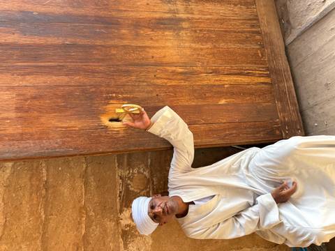 An Egyptian man in white galabeya points at a traditional wooden lock on a large rustic door.