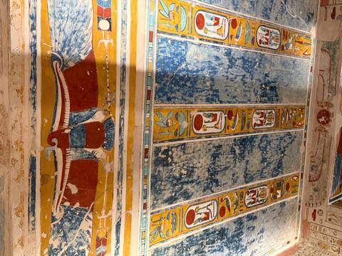 Vibrant painted ceiling relief with a winged sun disk and hieroglyphic bands in an ancient Egyptian tomb.