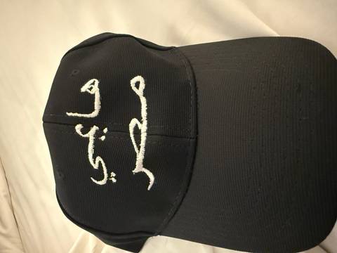 Close-up of a black baseball cap embroidered with Arabic script on a neutral background.