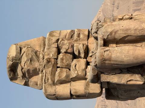 A partially ruined colossal stone statue against a clear sky, likely one of the Colossi of Memnon.