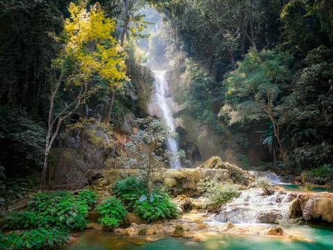 Multi-tiered Kuang Si waterfall cascades through dense jungle into turquoise pools surrounded by greenery.