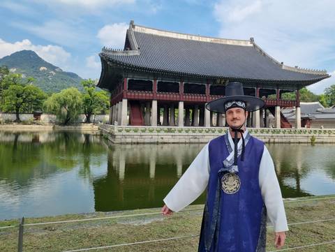 Man in traditional hanbok standing before Gyeongbokgung Palace pavilion reflected in a pond
