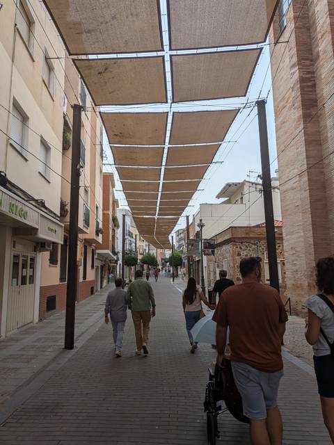 People stroll a narrow Spanish street shaded by canvas panels strung overhead between low buildings.