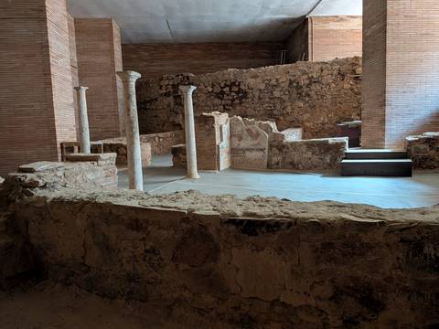 Exposed Roman-era stone walls, columns and mosaics displayed inside an archaeological site with soft indoor lighting.